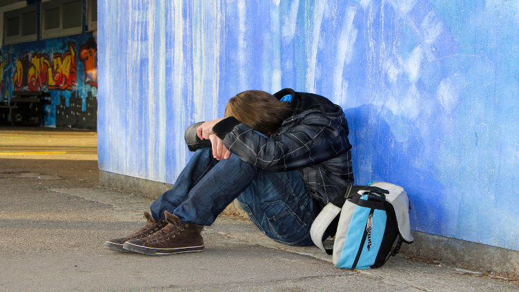 Zu sehen ist ein Jugendlicher, der vor einer Wand sitzt und seinen Kopf auf die Beine gelegt hat. Er sitzt in verschlossener Haltung da. - Auch digital ist brutal!