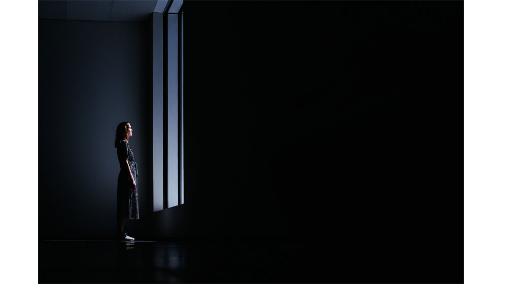 Zu sehen ist eine Frau, umgeben von Dunkelheit, die vor einem Fenster steht. Hell angestrahlt vom hereinfallenden Licht.